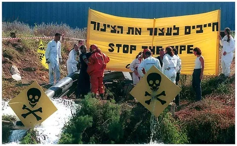 פעילים בחליפות מגן מפגינים ליד צינור שפכים, מתחת לשלט ״חייבים לעצור את הצינור״, ולפניהם שלטי אזהרה מחומר רעיל.